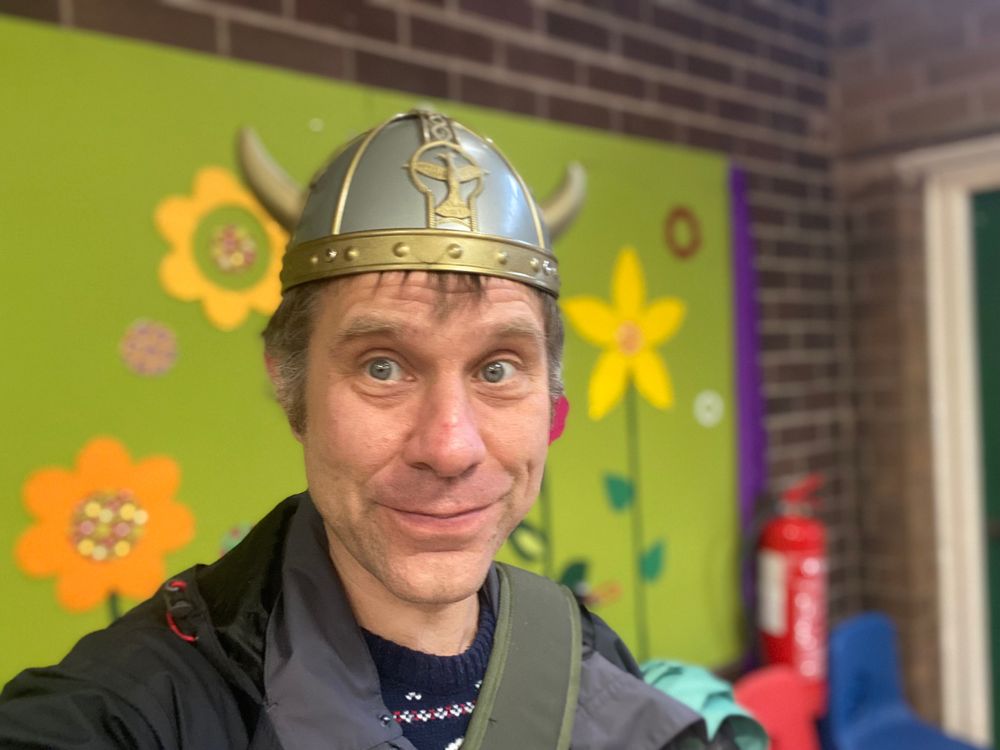 Selfie. I am wearing a plastic Viking helmet. Background: a brightly coloured wall display with yellow flowers on a green background.