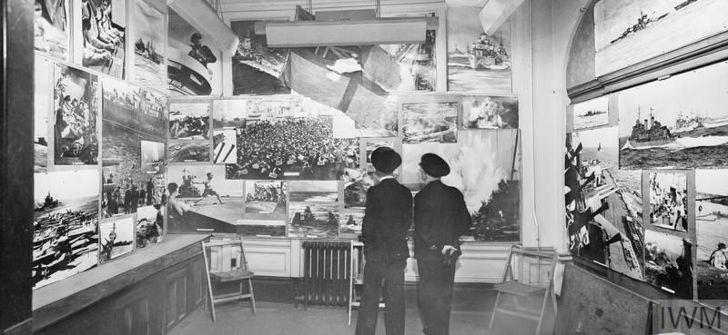 Two men in naval uniform look at photographs n the walls of a room. In the centre, and above the men, is a flag.