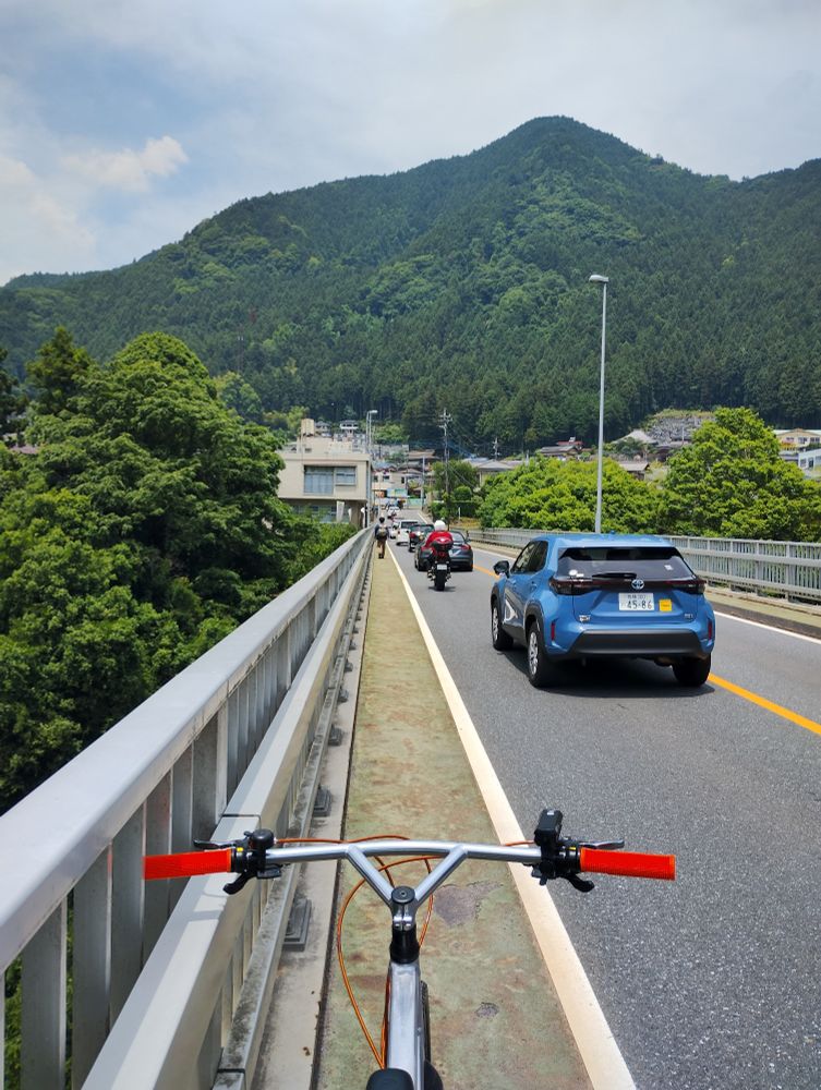 The front of a silver bicycle seen from the saddle. The bike is on a pavement with a road on the right. The road leads to lush green mountains. Cars on the road.