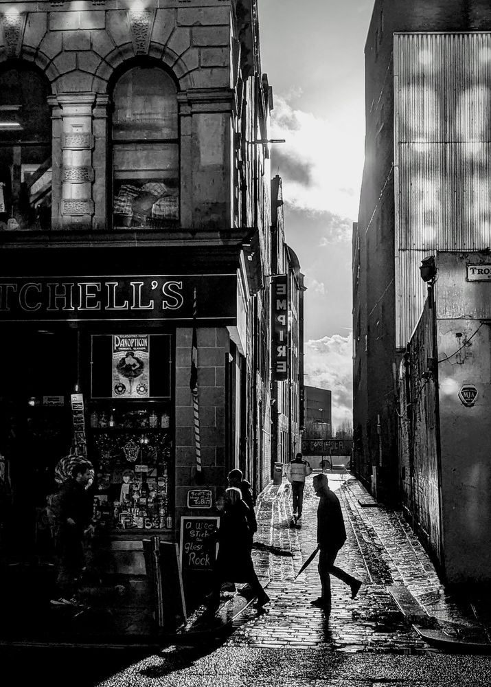 A black and white photograph of people on the Trongate, Glasgow, Saturday 25th Jan 2025. I just loved the sunlight reflecting from the buildings behind me. Made a lovely change from the storm shenanigans the previous day. Incidentally, the building on the left is the Britannia Panopticon music hall, the world's oldest surviving music hall and where in 1906, a 16 year old Stan Laurel made his stage debut.
