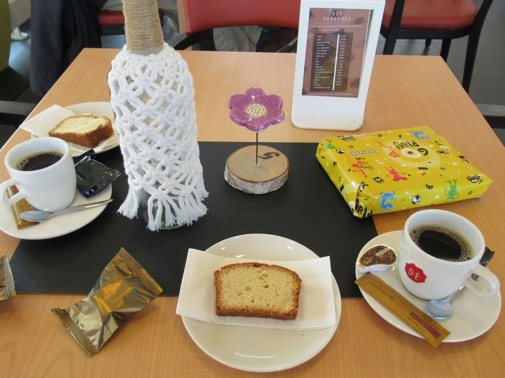 A picture from a table with on it two cup of coffee and two plates with a small cake piece on it. The table is decorated with a flower and a bottle. A small wrapped gift is laying on it to the side.