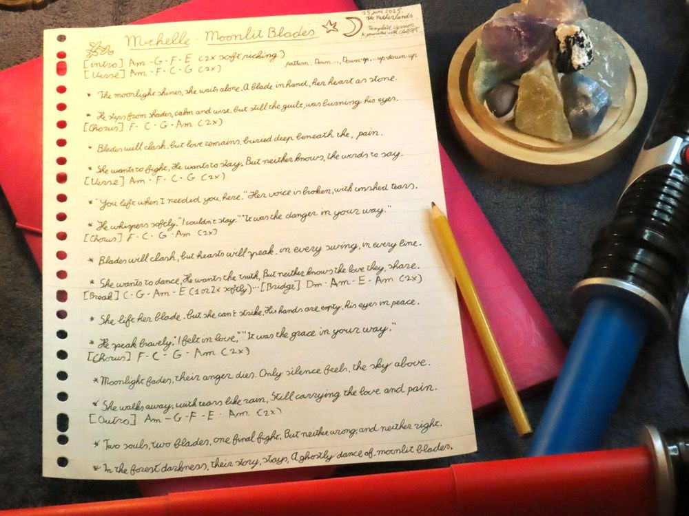 Picture of a sheet of paper with the song Moonlight Blades written out by hand. To the right sides next to the paper there is a pencil, a plastic blue light blade and different crystals and stones.