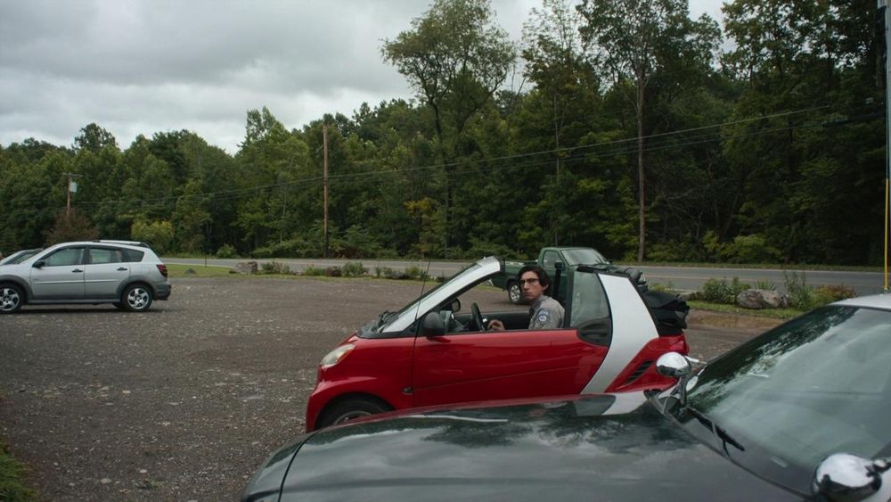 The fictional character Ronnie Peterson is driving in a red Smartcar. He's a character played by Adam Driver in the film, 'The Dead Don't Die'