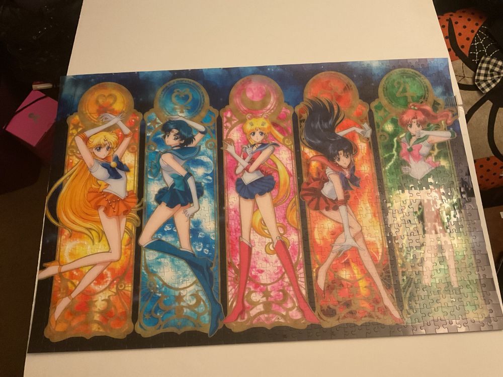 Image of a completed sailor moon puzzle. Puzzle is translucent plastic and has the five main sailor scouts, Venus, mercury, moon, mars, and Jupiter, posed in a stained glass window style with each in their own section. The puzzle is sitting on a foam core board in a slightly messy room. 