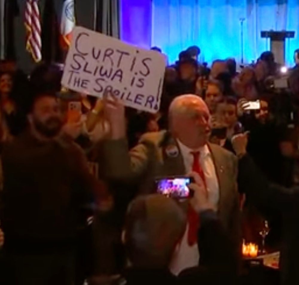Grainy screenshot of an old white man waving a handwritten sign reading "Curtis Sliwa is the Spoiler!"