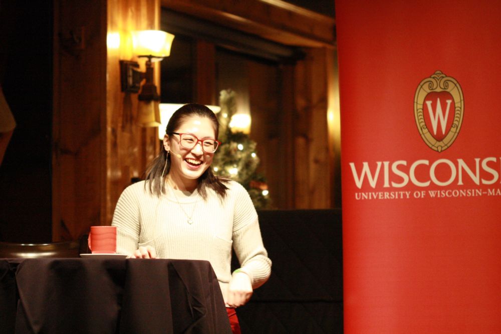 Edna Chiang giving a talk on stage. Edna is wearing red glasses and a gray sweater. In the background is a "University of Wisconsin-Madison" poster.