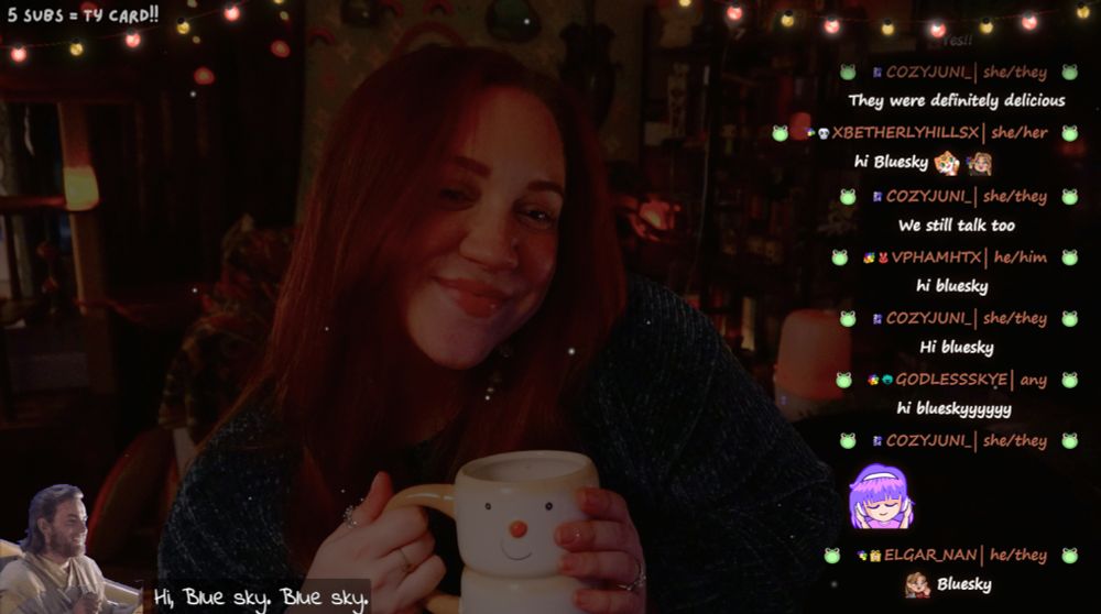 The image shows Ashley in a cozy well-decorated room illuminated by warm, ambient lighting. She is smiling while holding a snowman mug, seated at a desk with her hair down. The background is decorated with shelves full of books, candles, cat toys, and string lights.

On the right, a live chat is visible with friendly messages from viewers, saying hi to "Blue Sky."  At the bottom, text repeats, "blue sky" and there is a small graphic of obi wan