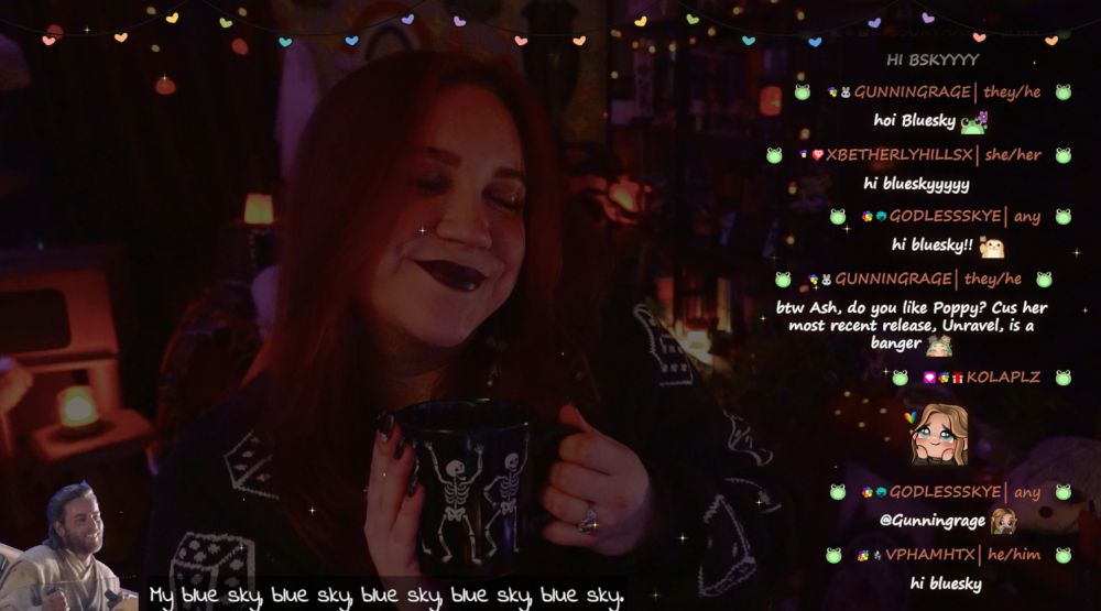 The image shows Ashley in a cozy well-decorated room illuminated by warm, ambient lighting. She is smiling while holding a black cat mug, seated at a desk with her hair down. The background is decorated with shelves full of books, candles, cat toys, and string lights.

On the right, a live chat is visible with friendly messages from viewers, saying hi to "Blue Sky."  At the bottom, text repeats, "blue sky" and there is a small graphic of obi wan