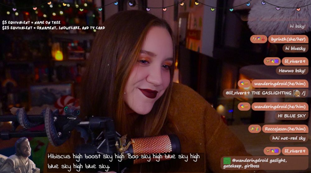 This image shows Ashley smiling warmly, sitting in a cozy environment with warm lighting and holiday decorations in the background.
At the top: "$5 equivalent = Name on tree | $25 equivalent = Ornament, snowflake, and TY card" with colorful heart-shaped lights as decoration.
On the right: A chat overlay from a livestream with messages from viewers, including phrases like "Hi bluesky," and mentions of the streamer's name, pronouns, and lighthearted comments.
At the bottom left: A dragon toy and a small image of Obi Wan Kenobi sitting.
A caption at the bottom reads: "hibiscus high boost sky high boo sky high blue sky high blue sky"