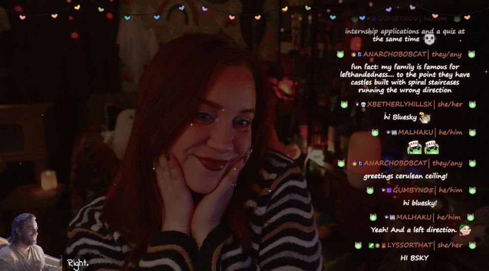 The image shows Ashley in a cozy well-decorated room illuminated by warm, ambient lighting. She is smiling, with her hands on her cheeks, seated at a desk with her hair down. The background is decorated with shelves full of books, candles, cat toys, and string lights.

On the right, a live chat is visible with friendly messages from viewers, saying hi to "Blue Sky."  At the bottom, there's text that says "right" and there is a small graphic of obi wan