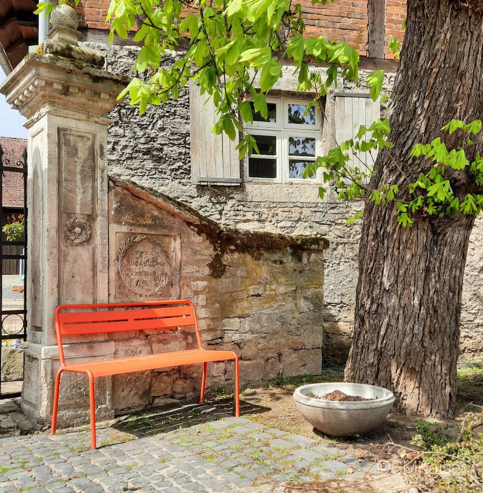Orange-rote Bank steht neben der Toreinfahrt zu einem Bauernhaus, rechts ein Baumstamm, Mitte ein Fenster hinter einer Mauer.
