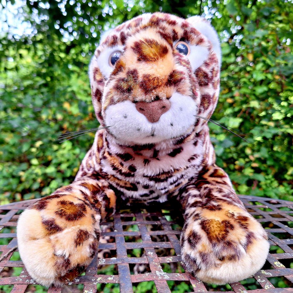 Plüschleopard, der sich mit zwei Pfoten auf dem eisernen Geflecht der Gartentischplatte festhält. Dahinter grüne Büsche.