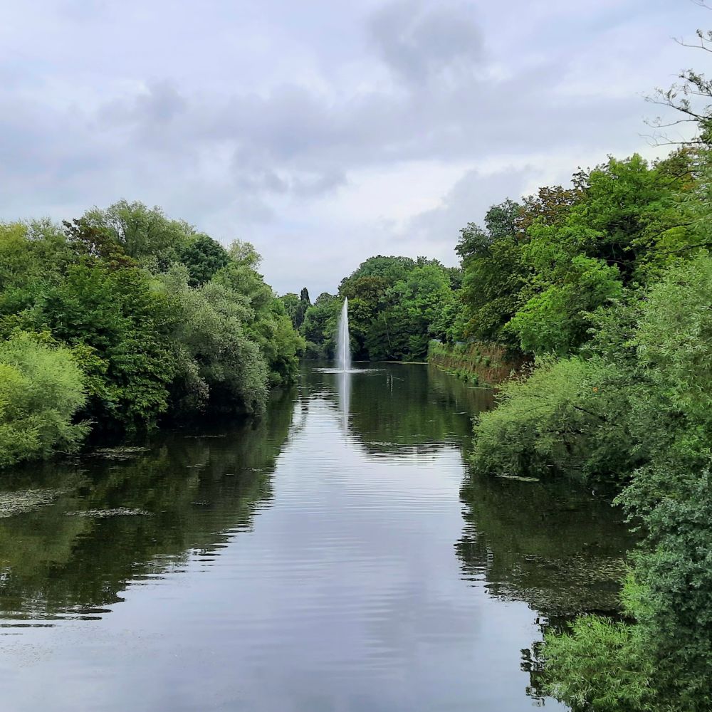 Die Saar, links und rechts mit Büchen und Bäumen bewachsenen Ufer, mittig eine Wasserfontäne mitten im Fluss.