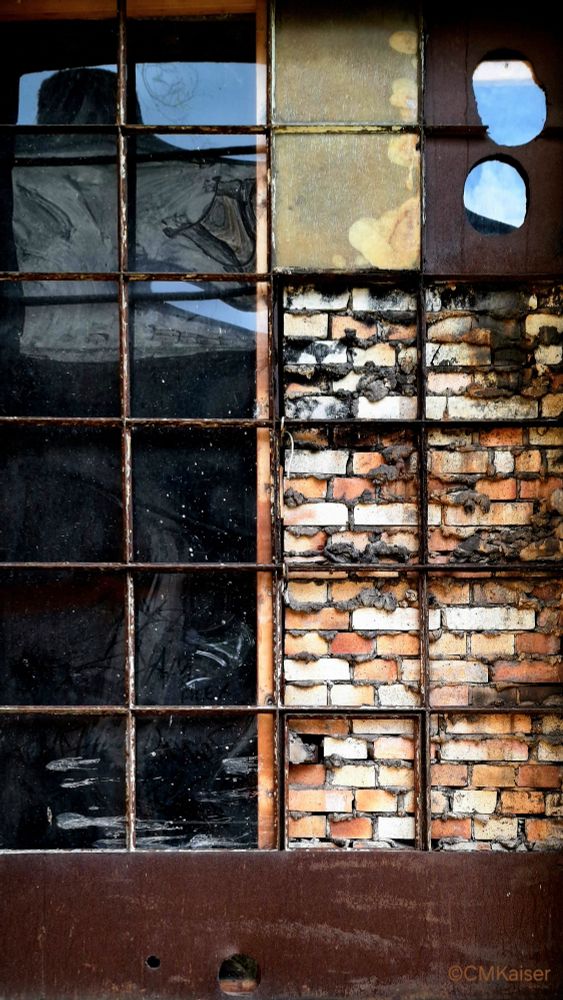 Verrostetes Sprossenfenster, linke Seite mit spiegelnden Glasscheiben, rechte Seite zum Teil hintermauert mit Ziegelsteinen, im oberen Bereich verklebt bzw. mit verrosteter Eisenplatte geschlossen, in der sich Löcher befinden, so dass sich der blaue Himmel darin spiegelt.