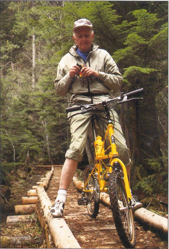 Color photo of Alex Moulton, in a casual outfit, standing astride a yellow small-wheeled mountain bike on a log bridge in a forest.
