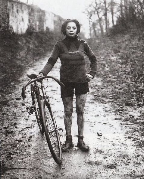 A woman, her legs splattered in mud, stands on a muddy path with her bicycle.