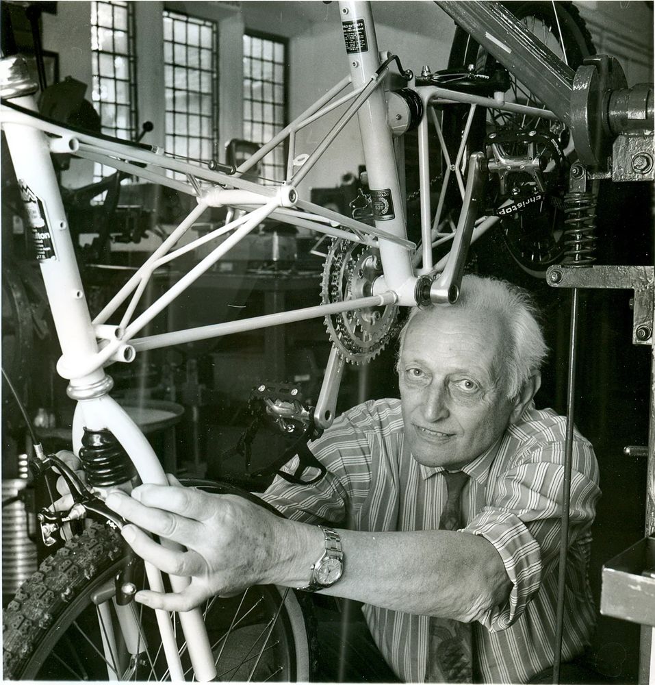 Black and white photo of Alex Moulton, in a shirt and tie, working on the front end of a bicycle. The bicycle is on a stand and Moulton is underneath it.