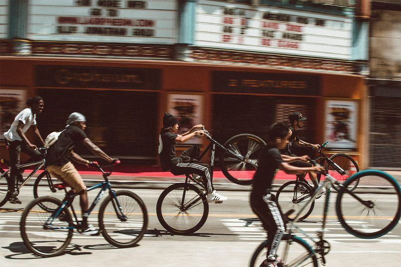 Photo of five kids on bikes riding on a city street. Three of them are doing wheelies. There's a movie marquee behind them.