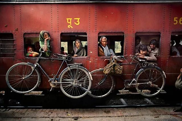 Photo of a train carriage, off of which two bicycles are hanging. Several passengers are visible through the open windows.