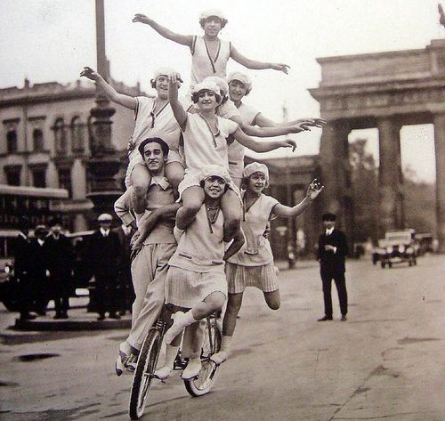 Photo of seven people riding a single bicycle on a city street. Many of them have outstretched arms. 