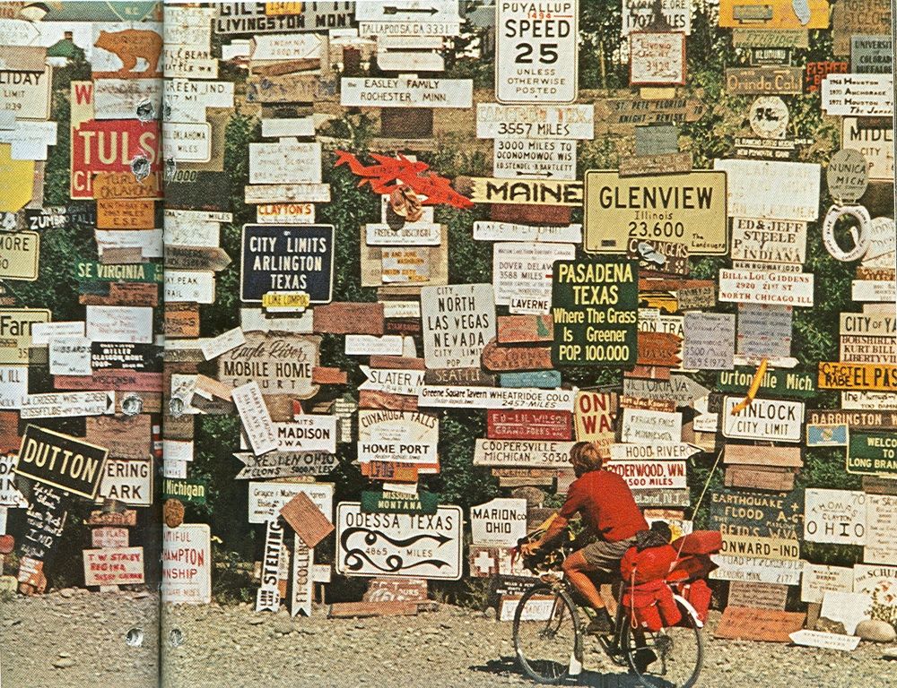 Photo of a man riding a fully-laden touring bike, complete with safety flag. In the background is a wall covered in road signs, many with the names of U.S. towns on them.