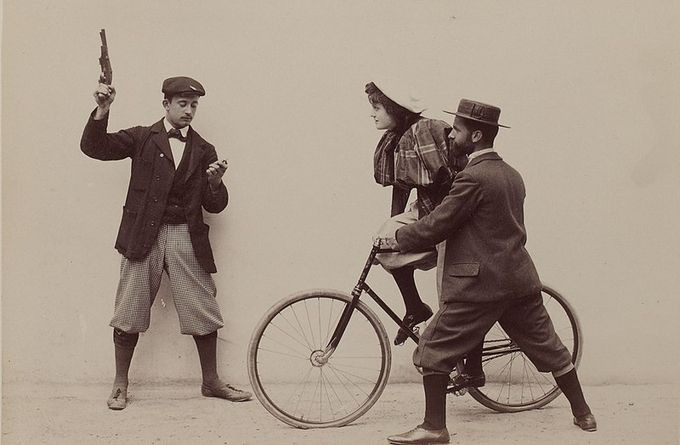 Photograph of a woman on a bicycle being held steady by a man, and another man holding a timer in one hand and pointing a gun into the air with the other. The caption says this photo takes place at the Buffalo Velodrome in Paris, but it looks like a studio photograph, because the background is completely blank.
