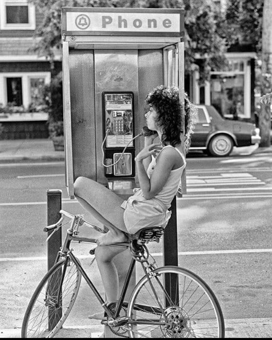 A young woman balances on a bike while talking on a payphone.