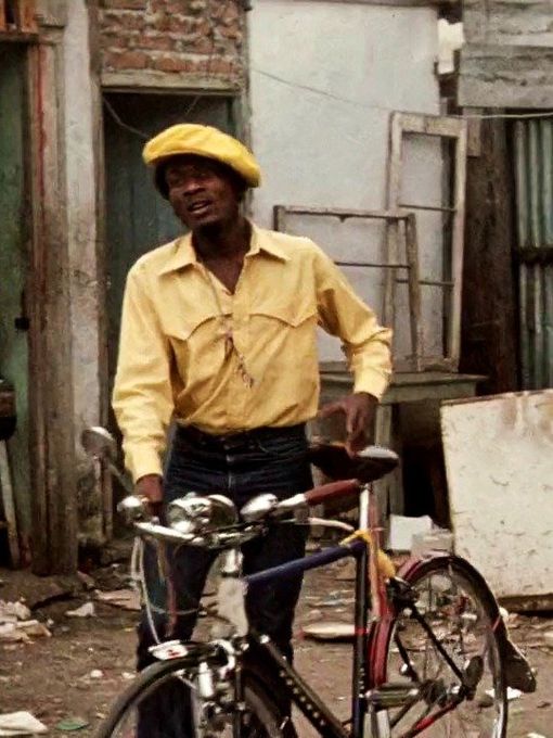 A man in a flat cap walks with a bicycle on a cluttered street.