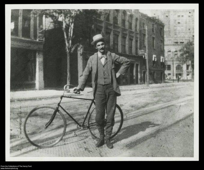 Antique photo of a man in a suit and hat standing with a bicycle.