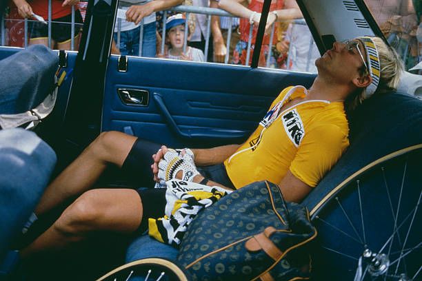 Photo of a man in a yellow jersey leaning back with his eyes closed in the back seat of a car. Two bicycle wheels are visible. Outside of the car you can see a crowd of people behind a barricade.