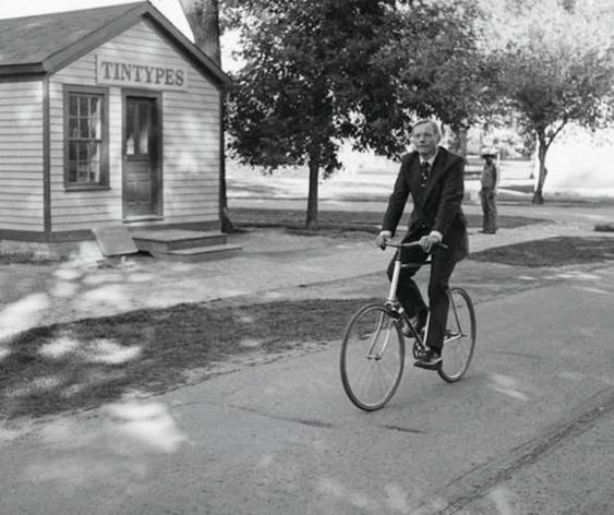 Photo of a man in a suit riding a bicycle past a small house with a sign on it that says, "Tintypes".