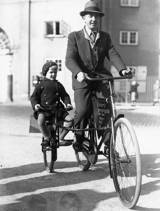 Man and boy riding side-by-side via something like a Trail-a-bike but it connects to the down tube rather than the seat post.