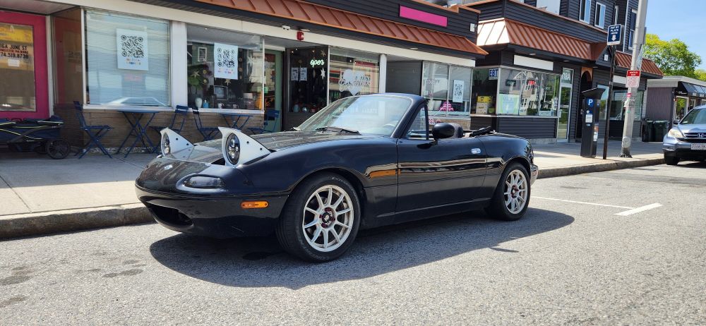 Two glossy white boxed pop-up headlights on a black convertible with 10 spoke glossy white wheels. The world's most popular and customizable sports car: The Mazda Miata. While This one is the last iteration of the first generation of the car, a 1996. Having a naturally aspirated 1.8 liter inline four engine; it was offered turbo as well.