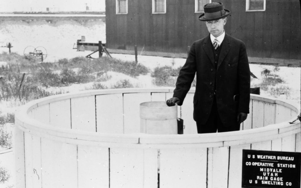 alt-text via GPT — "A man in formal dark coat and hat stands in a circular wooden weather monitoring station, with a sign reading "U.S Weather Bureau Co Operative Station Midvale Utah Rain Gage U.S Smelting Co" visible in this black and white historical photograph."