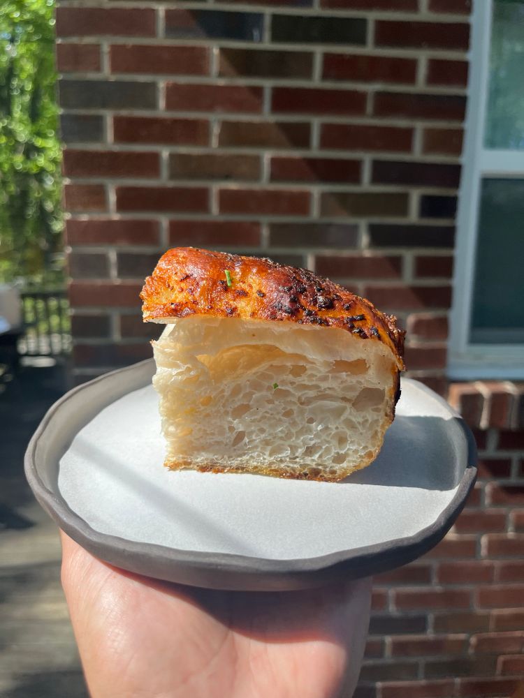 a picture of a slice of focaccia on a grey plate. I’m holding it up in the air with bricks in the background 