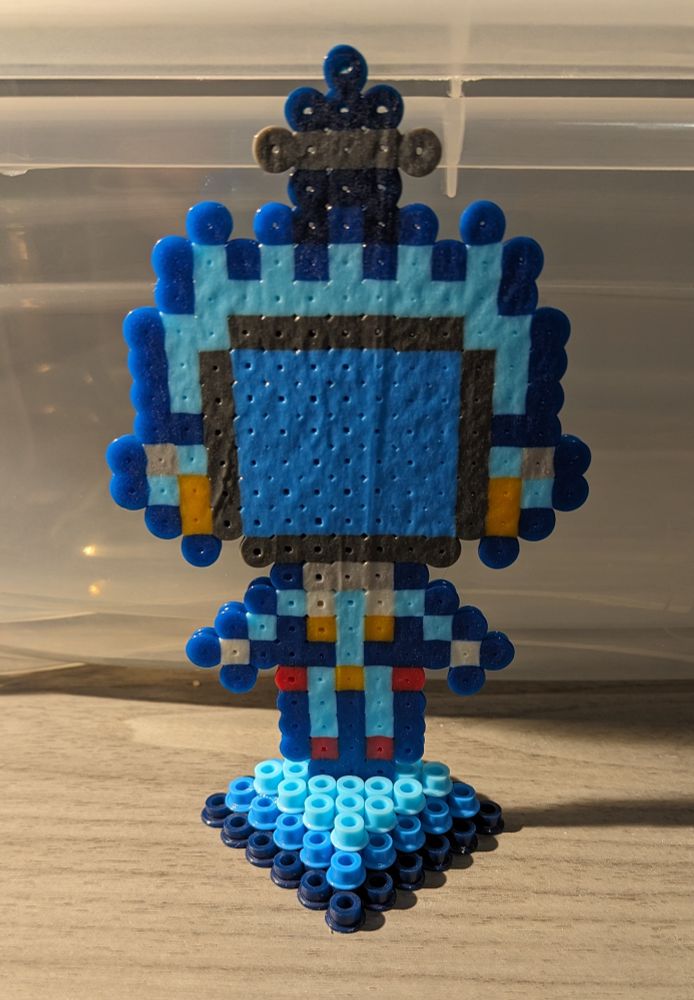 A photo showing a standee of a pixelated Canti, a TV-headed robot from the anime FLCL. The standee itself is made of perler beads, i.e. plastic beads that can be arranged into patterns and then ironed to fuse/melt them together into flat objects.