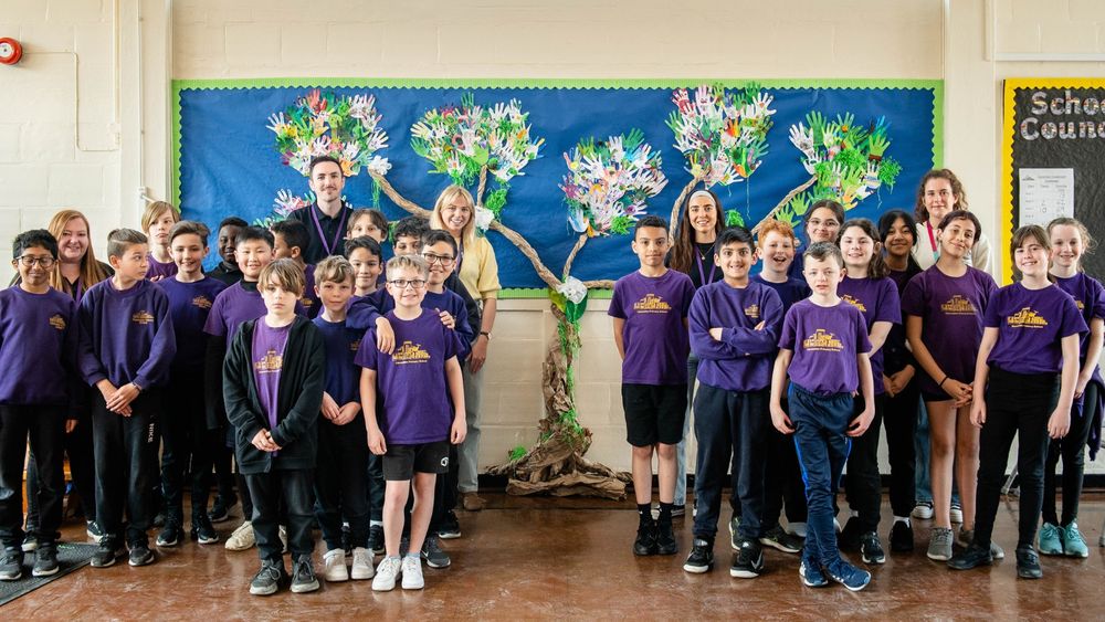 Group photo of students and Computing Ambassadors in a school hall with colorful artwork on the wall, all wearing purple school uniforms.