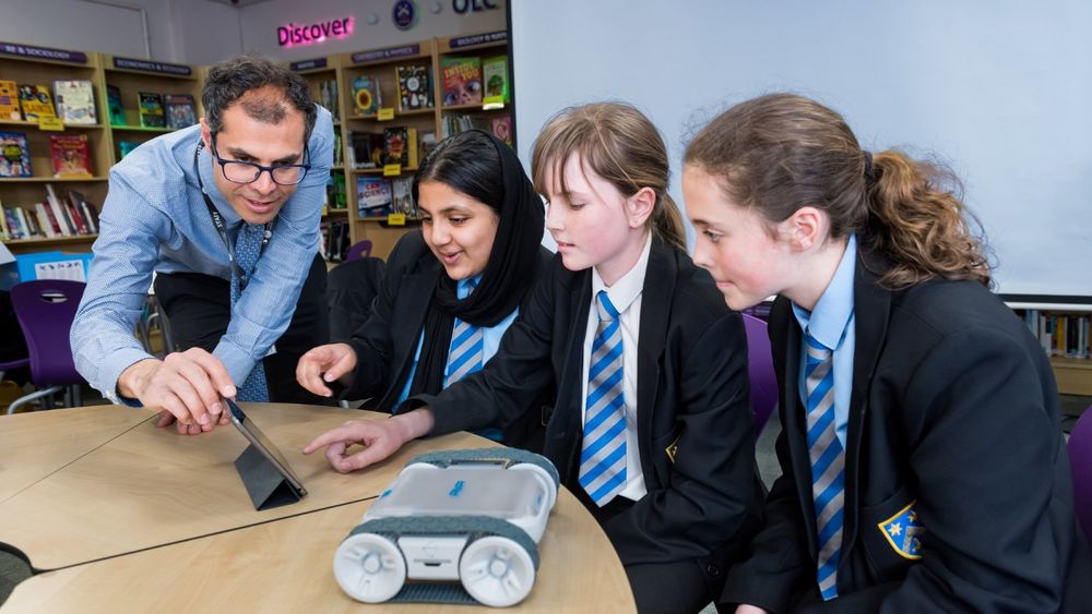 A teacher and three students in school uniforms engage with a tablet and a small robot on a table in a library setting.