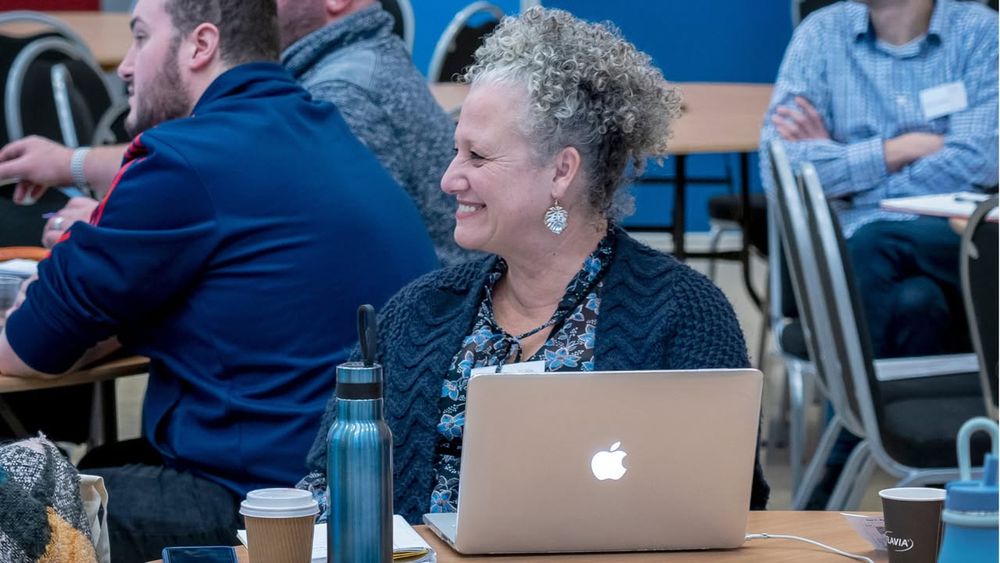 A person with curly hair smiles while using a MacBook at a busy computer science CPD course. Several attendees are visible in the background.