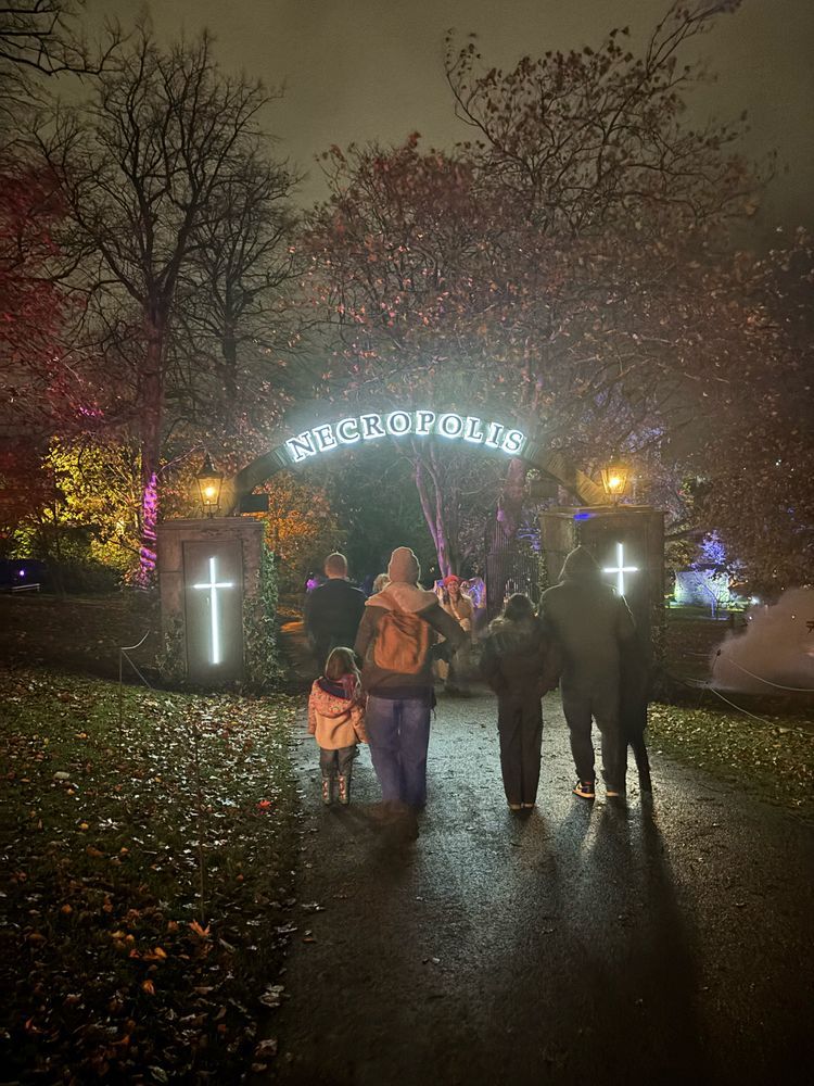 A group of people walk towards a gate with the word NECROPOLIS illuminated in neon above their heads. There are illuminated crosses either side.