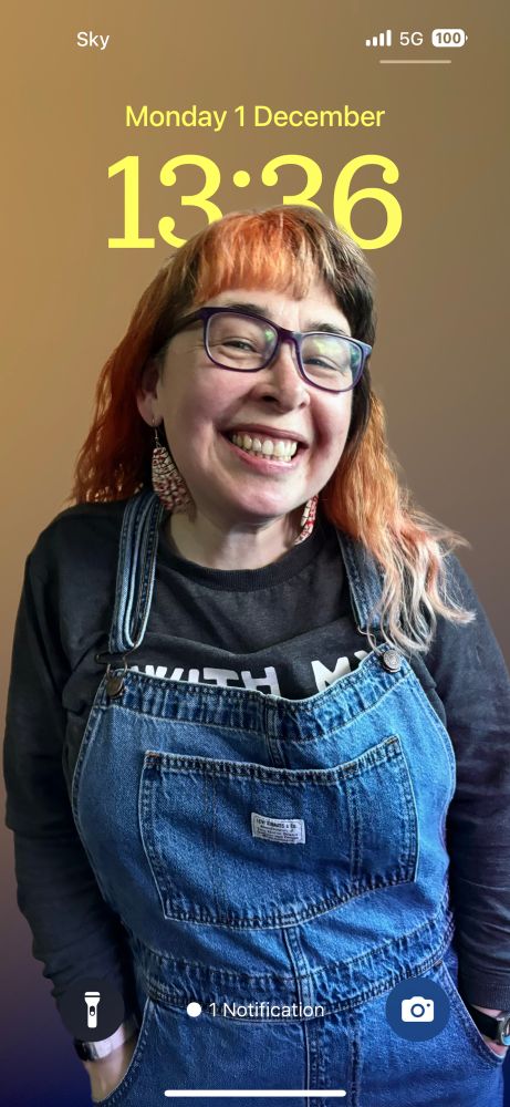 A young woman in dungarees and a black t-shirt smiles out at the camera. Her long hair is coloured orange and black and she wears glasses. The picture is being used as a phone background screen and behind her it shows the date and time at the point the screenshot was taken. 