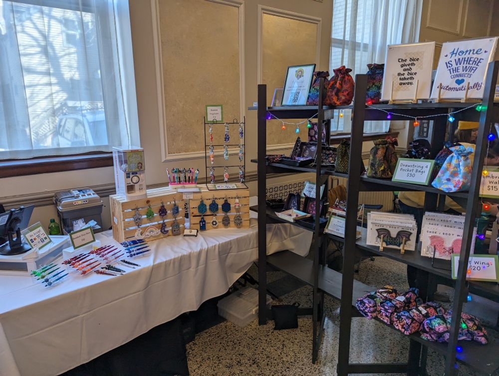 A photo of a booth at a craft fair, with a long white table with displays of keychains and a dark brown shelving unit with dice bags and dice trays