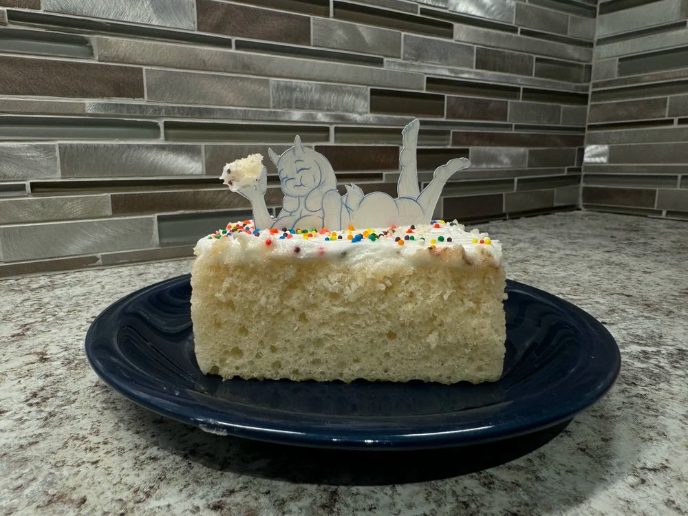 A paper cut out doll of Toriel is laying on top of a piece of cake and eating it. 