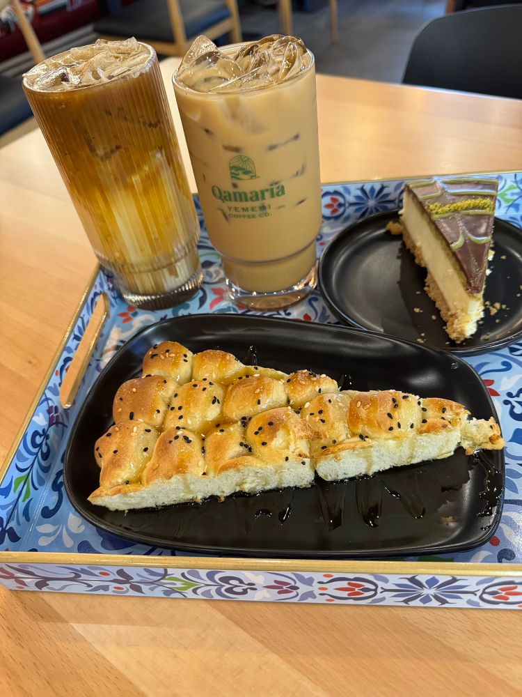 A photo of a tray holding two iced coffees, a pastry, and a slice of cheesecake, all served on a wooden table.
The latte  on the right has the text "Qamaria Yemeni Coffee Co." printed on the side. Both drinks appear to be layered, with milk/cream at the bottom and coffee on top, and are filled with ice.
 * Pastry: A triangular-shaped, pull-apart pastry is on a black plate in the foreground. It is golden brown, topped with black sesame seeds, and drizzled with a dark syrup or honey.
 * Dessert: A slice of cheesecake is on a smaller black plate to the right. It has a graham cracker crust, a cream-colored filling, and is topped with a dark chocolate layer and a garnish of what appears to be chopped pistachios.
🖼️
