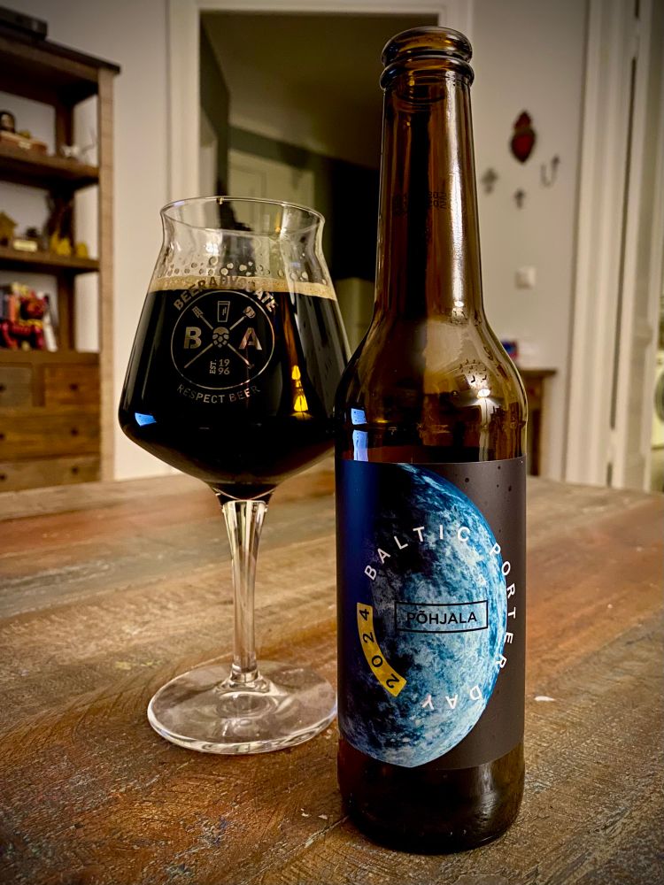 A bottle of Baltic Porter (Day 2024) beer from Põhjala is next to a Teku glass on a wooden table. The beer is dark, and the bottle label features a design with a planet motif. The setting appears cozy with a blurred background.