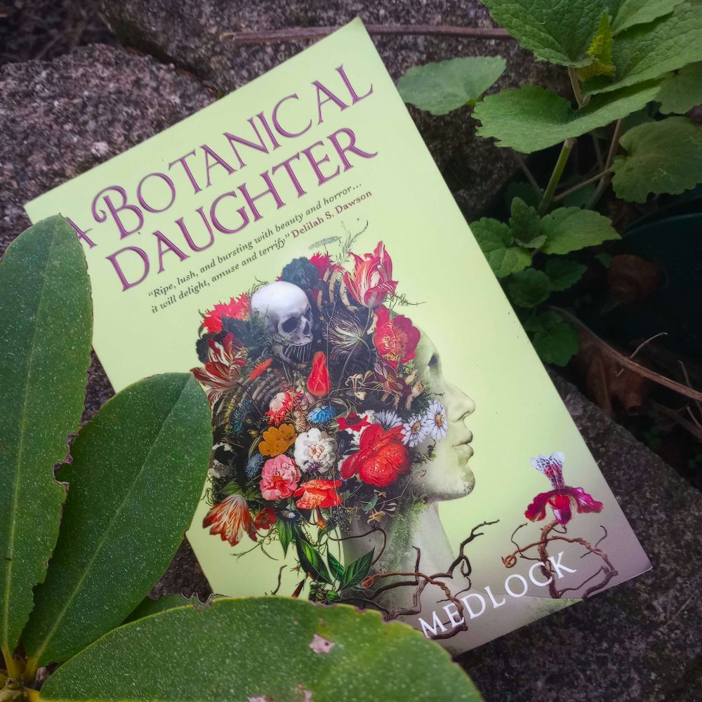 a paperback of Noah Medlock's A Botanical Daughter lying on a stone ledge, surrounded by greenery. the cover is pale green with a central image of a girl in profile made from flowers and fungi, with a skull in her floral hair