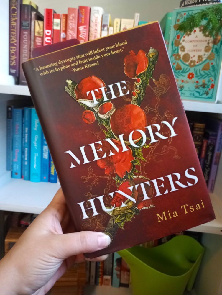 hardcover of Mia Tsai's The Memory Hunters, held up against a rainbow shelf. The cover is dark red, with paler red fungi growing on branches and the faint outline of branches and fungi that are no longer there