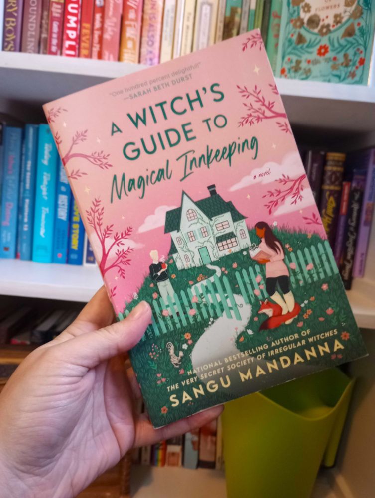 paperback of Sangu Mandanna's A Witch's Guide to Magical Innkeeping held up again a rainbow shelf. the cover is pink with a cottage on a green hill behind a fence, with a man behind the fence and a woman in front of the fence with a fox at her side and an undead rooster nearby