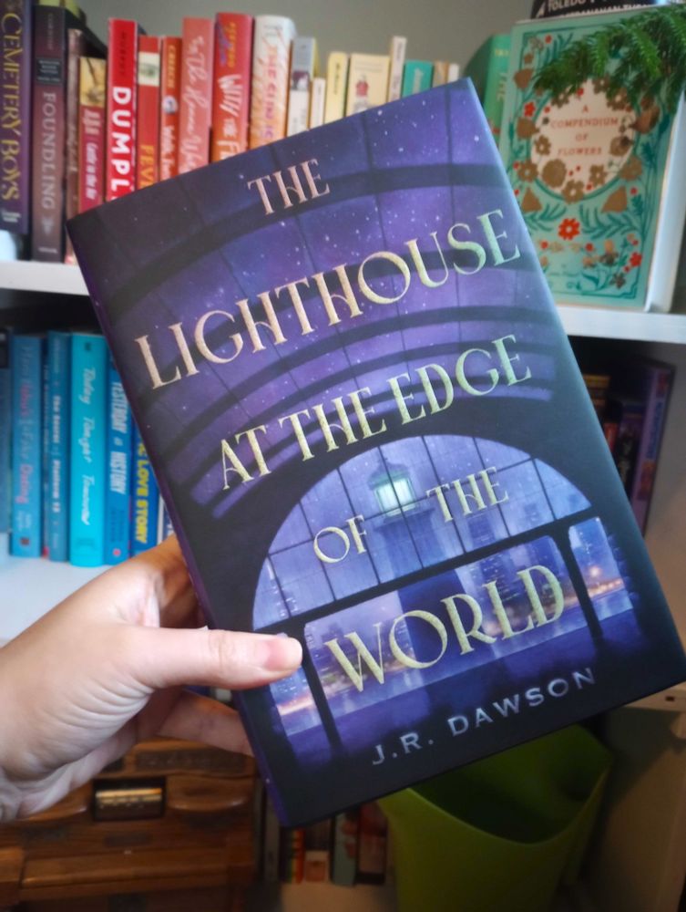 hardcover of J.R. Dawson's The Lighthouse at the Edge of the World, held up in front of a rainbow shelf. The cover is dark blue, a view of a lighthouse in the misty evening through a window and stars overhead through a skylight