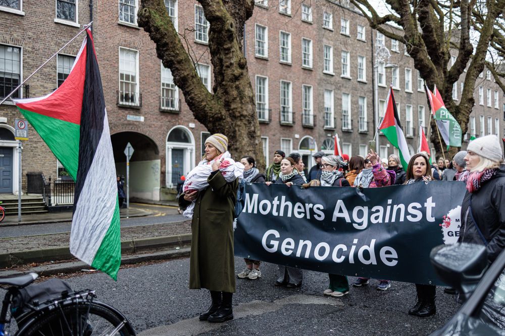 A woman stands in the road, holding something bundled in white sheets and stained with red, and emotional prop to represent all the children slain in Palestine.  She stands in front of a large black banner being carried by no fewer than six women.  In large white font the banner reads "Mothers Against Genocide".  Several of the protesters are carrying Palestinian flags.  Behind them two bare-leaf mature trees stretch tall out of frame, and behind the trees are rows of Georgian four-story red brick houses.